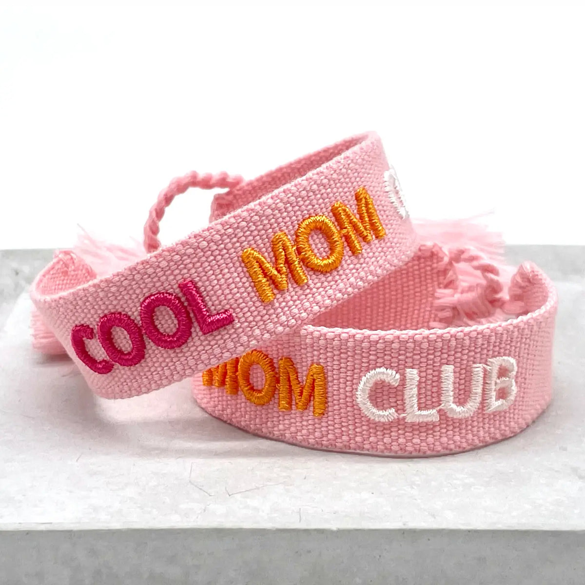Armband „COOL MOM CLUB" aus rosafarbenem Webstoff mit bunter Stickerei in Pink, Orange und Weiss, mit geflochtenen Fransen-Enden, auf einem Marmorstein drapiert – zwei Armbänder übereinandergelegt - el encanto