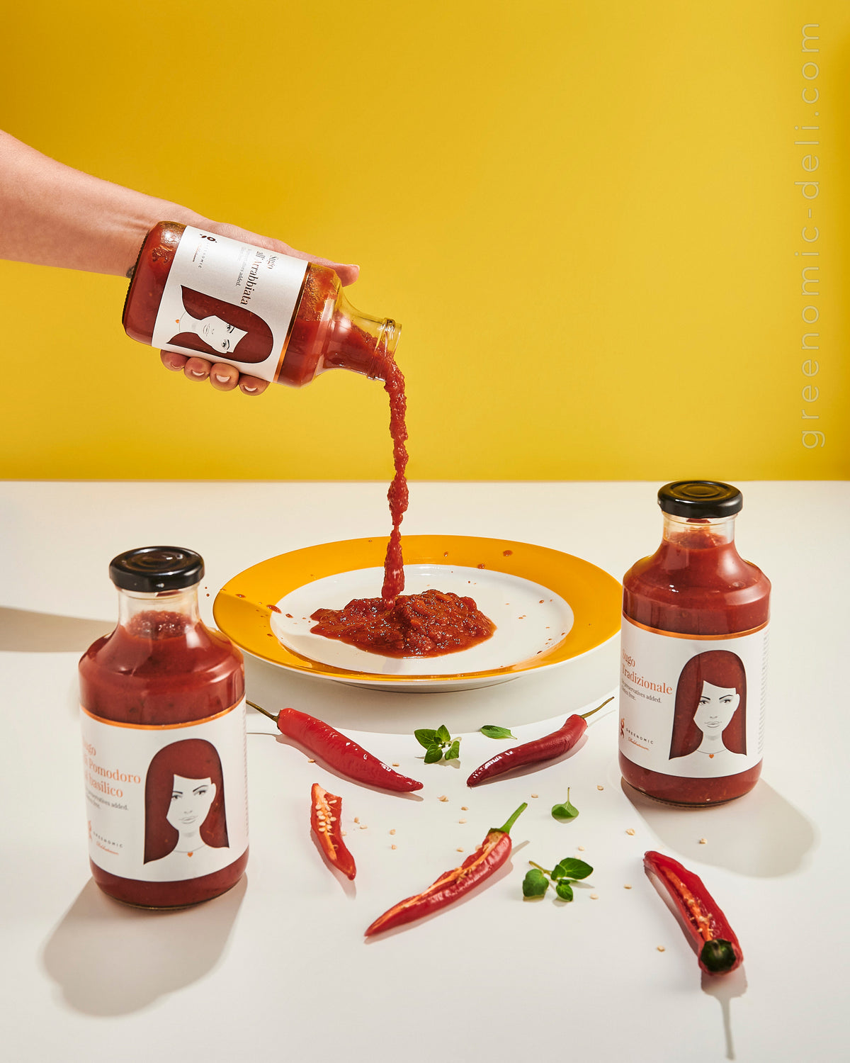 Greenomic Sugo Sortiment – Lifestyle Bild Hand Giesst Arrabbiata in Teller mit Chilis und Basilikum vor Gelbem Hintergrund – el encanto