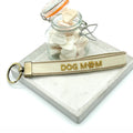 Schlüsselanhänger „DOG MOM" aus beigefarbenem Webband mit braunen Streifen und goldener Stickerei mit Pfoten-Icon, bronzefarbenem Schlüsselring, auf einem Marmorstein drapiert neben einem kleinen Glasgefäss mit weissen Steinchen auf weissem Hintergrund - el encanto